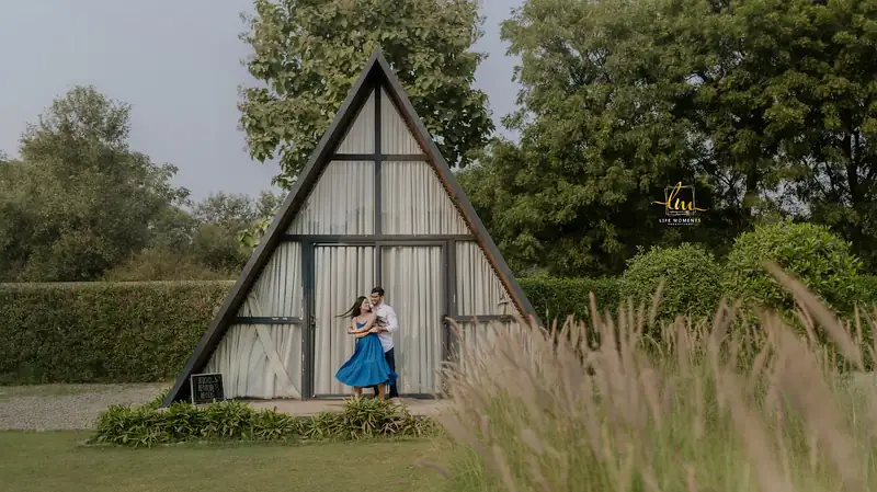 A couple standing in tall grass in front of a triangular wooden cabin.