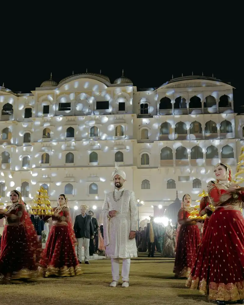 A groom walking in with dancers on either side, a lit venue facade behind him at night.