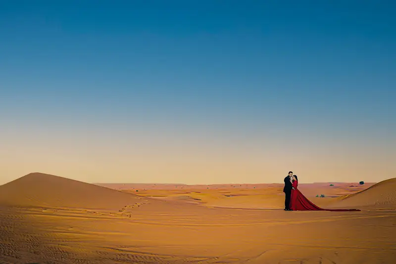 A couple standing alone on a sand dune at sunset, a long red train trailing behind them.