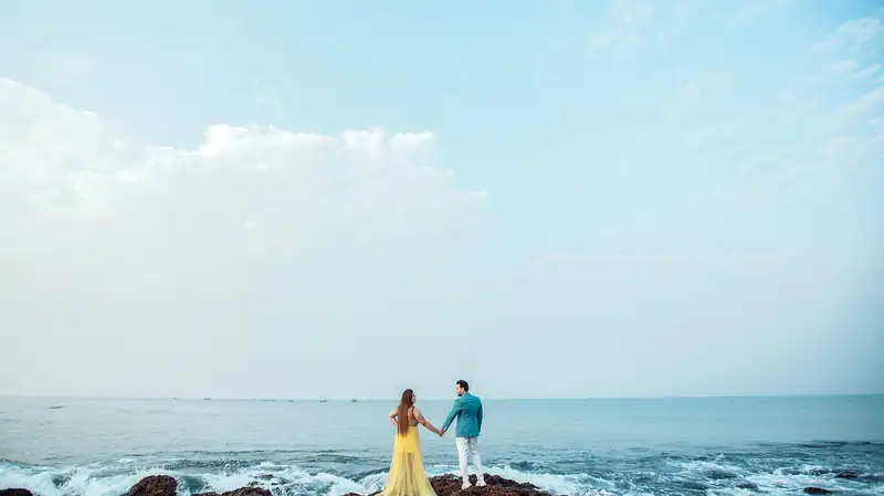 Two small figures holding hands on black rocks where the sea breaks, under an open sky.