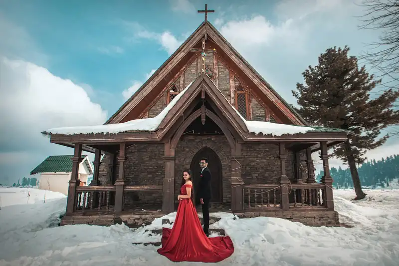 A wooden church under snow with a couple on its veranda, a red train spilling down the steps.